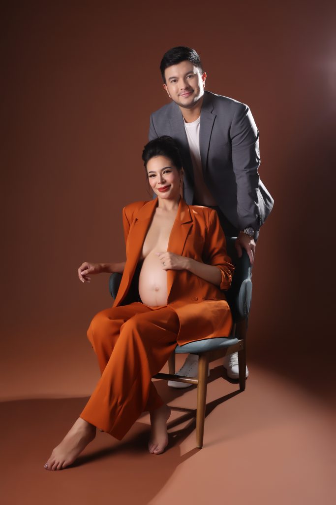 emma rueda ayala maternity photoshoot with brown background wearing an orange oversized suit with husband lucho ayala wearing a grey suit