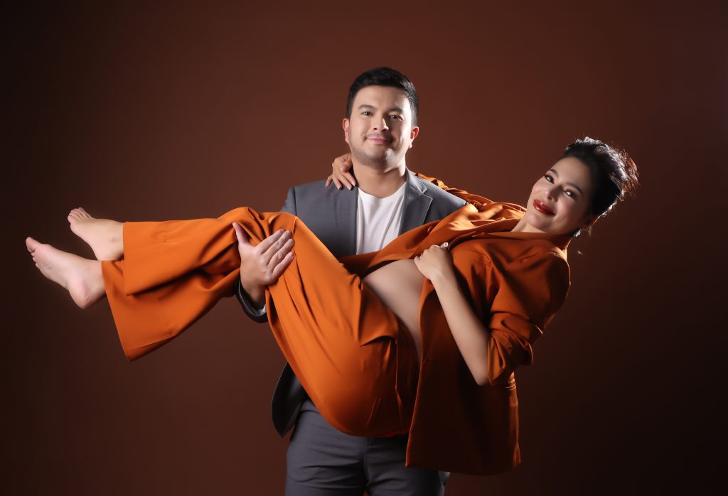 emma rueda ayala maternity photoshoot with brown background wearing an orange oversized suit with husband lucho ayala wearing a grey suit